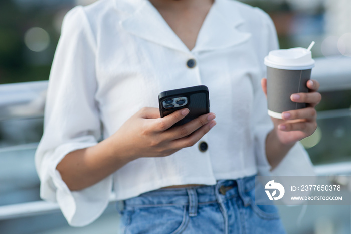 Cropped image of a young woman in a white dress holding a paper cup with coffee and a smartphone in hand in the city.