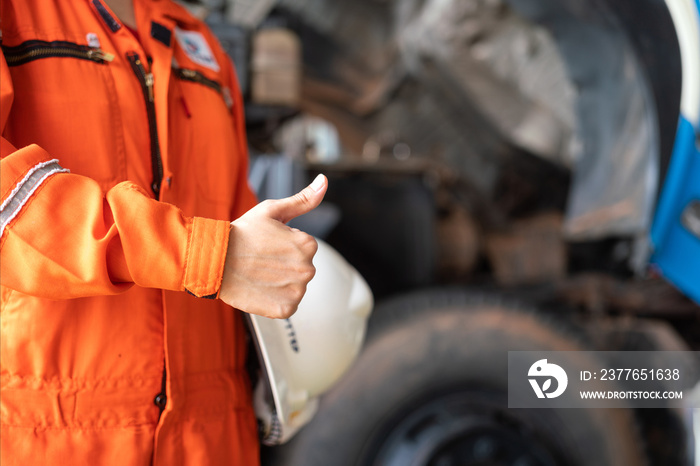 A repairman in orange coverall is thumbing up and holding white safety helmet with blurred background of garage workshop. Safe working practice in industrial concept.