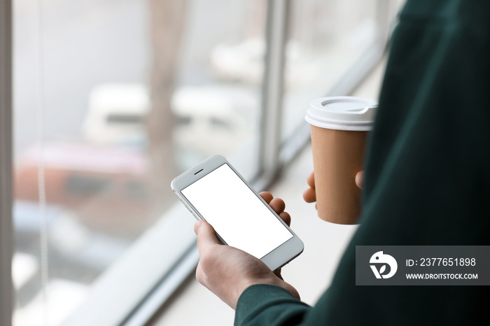 Young man with cup of coffee and modern mobile phone near window, closeup