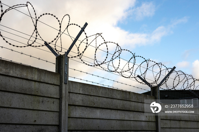 silhouette of security razor wire and barbed wire on top of a panel wall blue sky and clouds