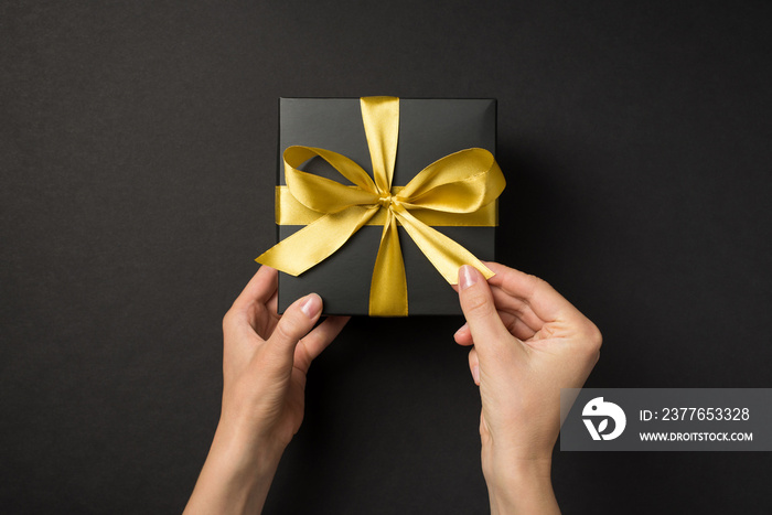 First person top view photo of hands opening black giftbox with golden satin ribbon bow on isolated black background