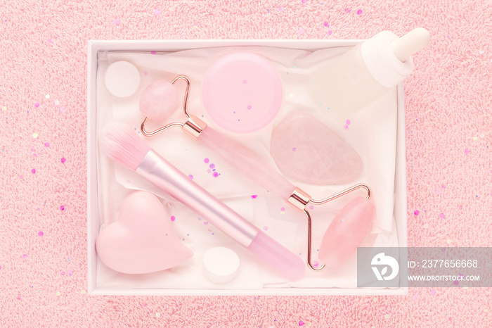 beauty box with rose quartz jade roller and massage stone, face serum and mask brush on pastel pink background