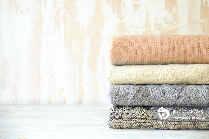 Bunch of knitted warm pastel color sweaters with different knitting patterns folded in stack on white wooden table, textured wall background. Fall winter season knitwear. Close up, copy space for text