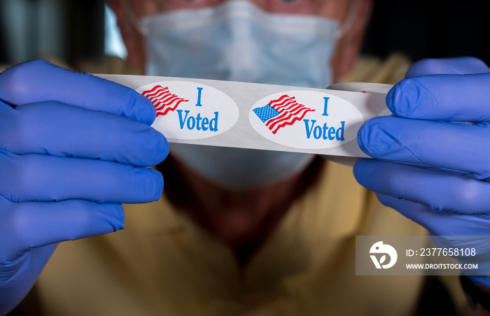 Election volunteer with face mask and medical gloves holding roll of I Voted buttons with USA Flag ready for in person voters in Presidential election