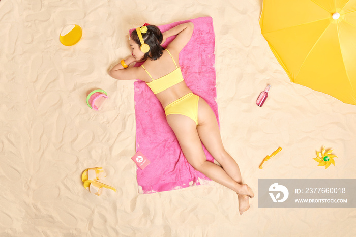 Relaxed Asian woman takes nap at beach listens serene music via headphones wears yellow bikini lies on pink towel sunbathes during sunny summer day. People leisure relaxation holidays concept