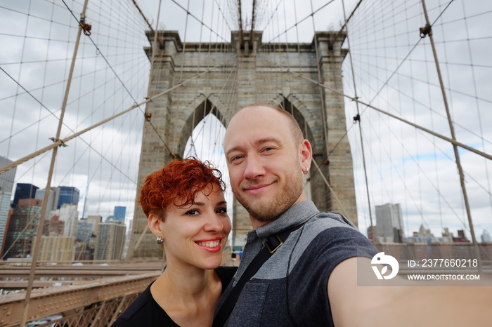 Happy selfie tourist woman and man taking self-portrait picture with smart phone on Brooklyn Bridge, New York City, Manhattan, USA. Young beautiful family couple travel, photographing NY landmarks.