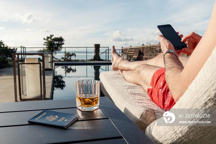 Man with alcoholic drink and passport relaxing next to swimming pool while using smart phone