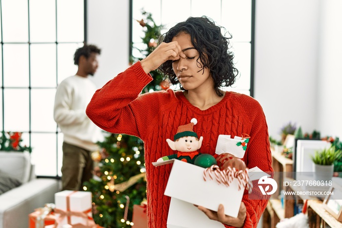 Young hispanic woman standing by christmas tree with decoration tired rubbing nose and eyes feeling fatigue and headache. stress and frustration concept.