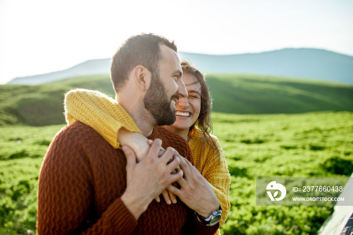 Lovely couple in bright sweaters hugging together on the green meadow, while traveling high in the mountains during the sunset