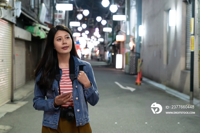 pretty asian girl tourist using navigation app on the phone while walking through shopping street in Osaka, japan at nighttime with neon lights at background