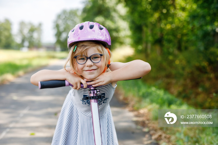 Portrait of active little preschool girl with glasses and helmet riding scooter on road in park outdoors on summer day. Seasonal child activity sport. Healthy childhood lifestyle