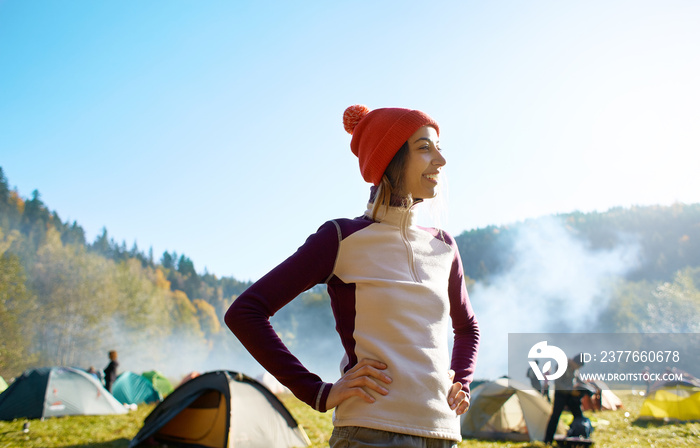 happy smiling woman in a red cap. Woman is standing in campsite on a meadow in the autumn forest on a bright sunny foggy morning