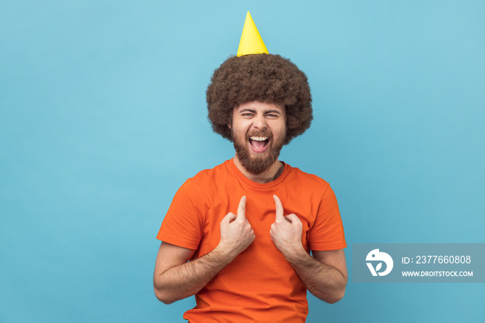 Portrait of crazy happy man with Afro hairstyle wearing orange T-shirt and party cone, looking at camera, pointing at himself, celebrating his birthday. Indoor studio shot isolated on blue background.