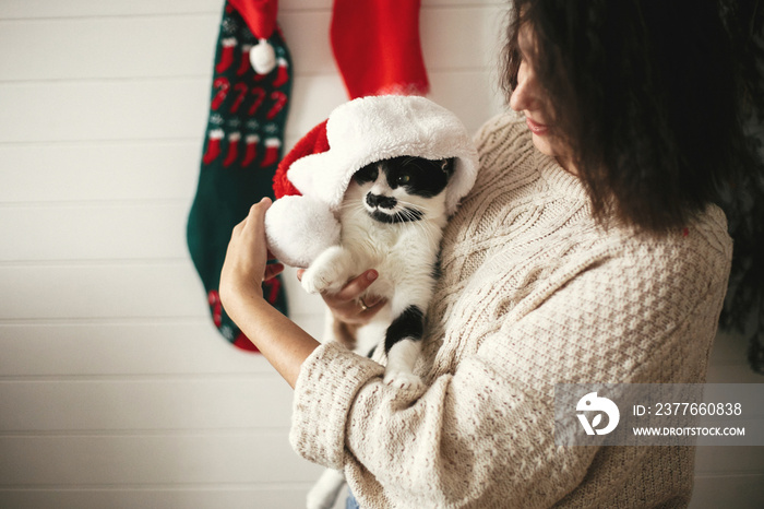 Stylish happy girl smiling and playing with cute cat in santa hat in background of  christmas tree lights and stockings. Young hipster woman in cozy sweater hugging kitty. Happy Holidays