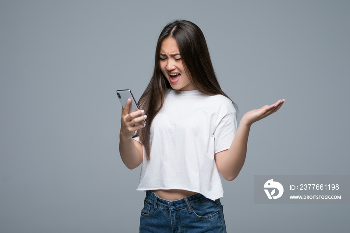 Angry mad young woman shouting and using mobile phone over gray background