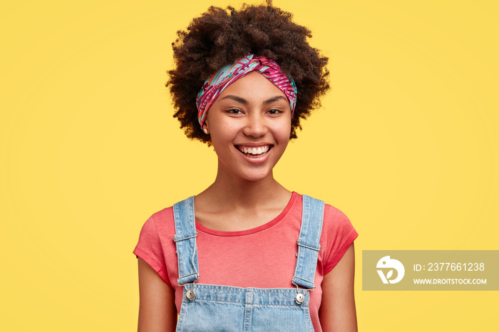 Happy pretty female housewife dressed in casual t shirt and denim overalls, has broad smile as did all work about house, poses against yellow background indoor. African American woman giggles