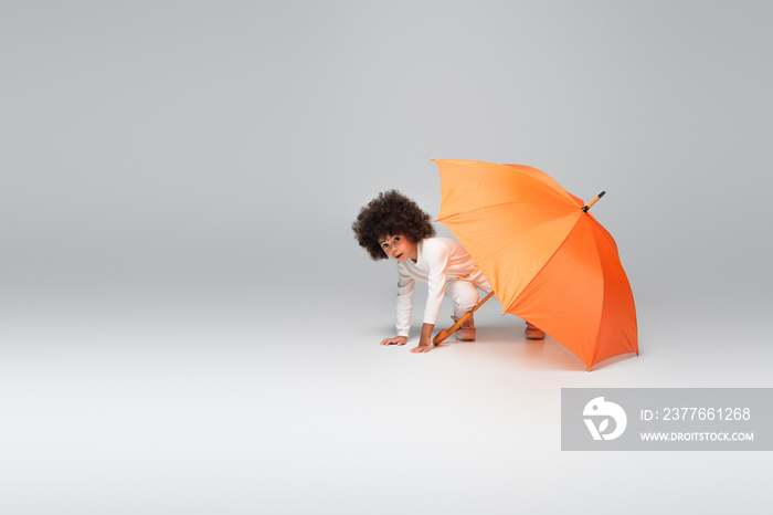 african american girl in white clothing looking at camera while crawling under orange umbrella on grey