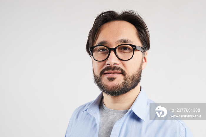 Portrait of content confident young Asian bearded man in glasses standing against light gray background