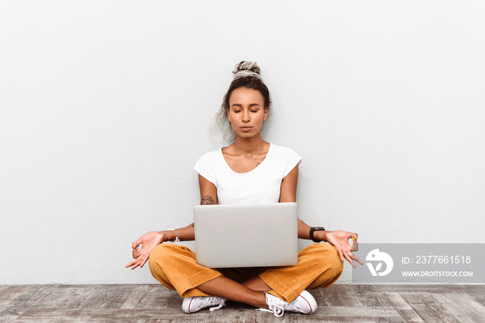 Concentrated young african woman with dreads sitting isolated over white wall background using laptop computer meditate keep calm.