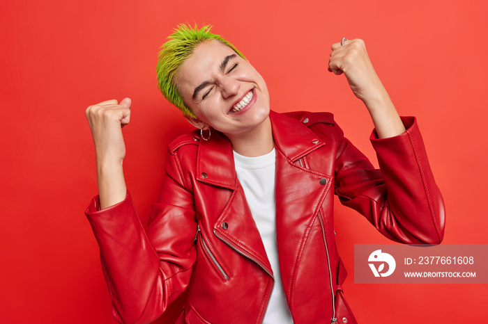 Overjoyed hipster girl with green hair makes yes gesture clenches fists celebrates success triumphs over something dressed in red leather jacket poses indoor wins prize smiles feels glad. Yes I did it