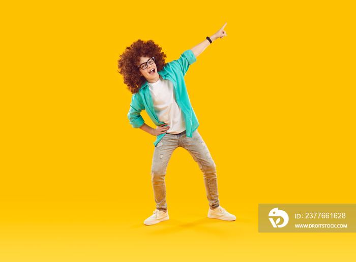 Happy child pointing away. Cheerful funny little boy in casual turquoise shirt, gray jeans, curly wig and glasses pointing aside to show something while standing on yellow color studio background