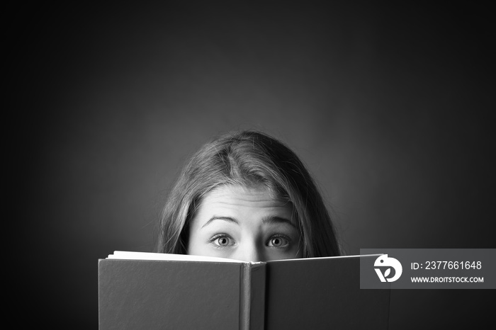 Black and white portrait of surprised teenage girl with book on dark background