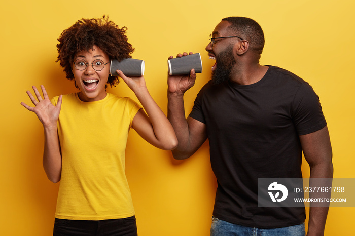 Happy dark skinned carries cup of takeaway coffee near opened mouth, pretends singing in microphone, amazed girlfriend holds paper mug near ear, listens carefully, gestures actively with palm