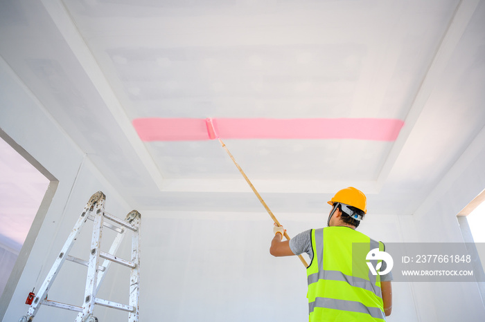 The painter is painting the ceiling inside the house or building. Use a white primer paint roller on the construction site.