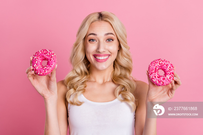 Portrait of attractive cheerful wavy-haired girl holding in hands two donuts isolated over pink pastel color background