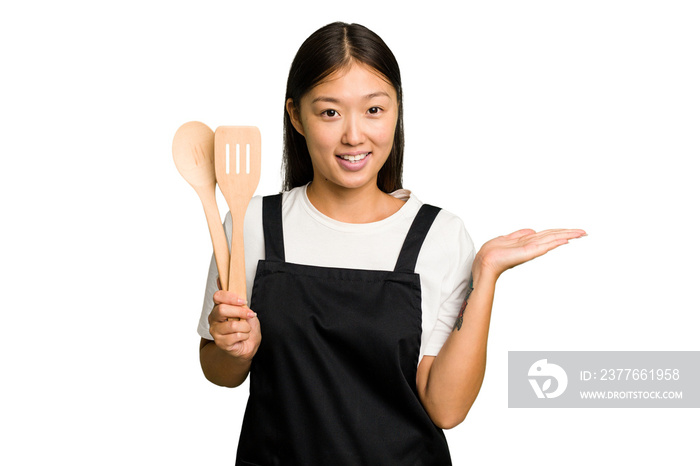 Young asian cook woman isolated showing a copy space on a palm and holding another hand on waist.