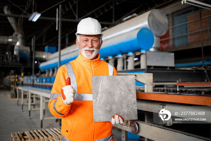 Factory production line worker standing by automated machine holding ceramic tile and thumbs up.