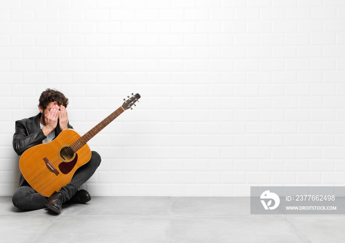 young musician man feeling sad, frustrated, nervous and depressed, covering face with both hands, crying with a guitar, rock and roll concept