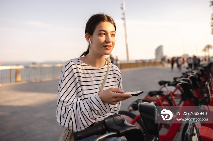 Calm young caucasian girl using bike sharing app on smartphone outdoors. Brunette looks away, wears casual sweatshirt. Lifestyle concept