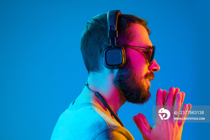 Enjoying his favorite music. Happy young stylish man in sunglasses with headphones listening and smiling while standing against blue neon background