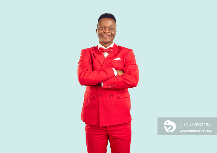 Male model in funky outfit posing in fashion studio. Happy smiling handsome young African American man wearing stylish red suit standing with his arms folded isolated on light background