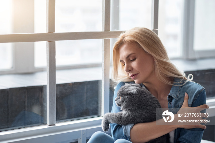 Blond woman holding cat near window.