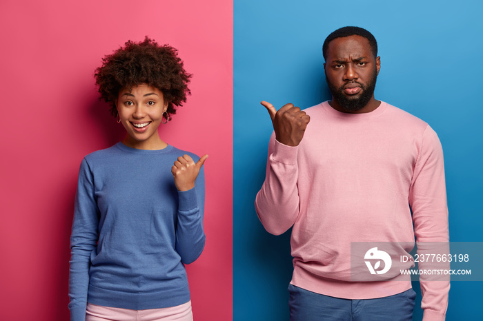 Positive woman and sullen displeased man point at each other, blame or suggest to choose him or her, wear blue and pink clothes, stand in studio indoor. Human facial expressions, advertisement