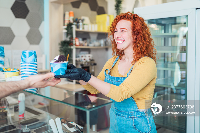 Beautiful and positive redhead ginger woman smiling and working in handmade ice cream store. She preparing and serving delicious sweet food.