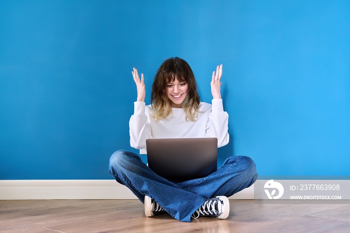 Emotional teenage female sitting on floor with laptop on blue background