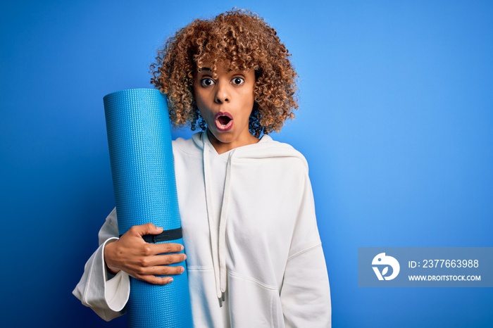 Young african american woman doing sport holding yoga mat over isolated blue background scared in shock with a surprise face, afraid and excited with fear expression