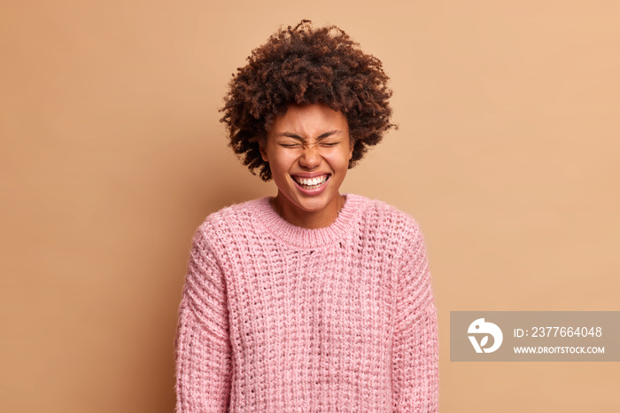 Amused carefree African American woman closes eyes and smiles broadly expresses pure emotions speaks to funny friend wears warm knitted sweater poses against beige background laughs out loudly