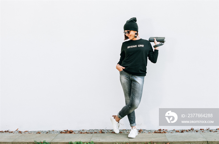 Woman in black outfit with bright accessories stands near wall. Black sweatshirt with Shine On golden print