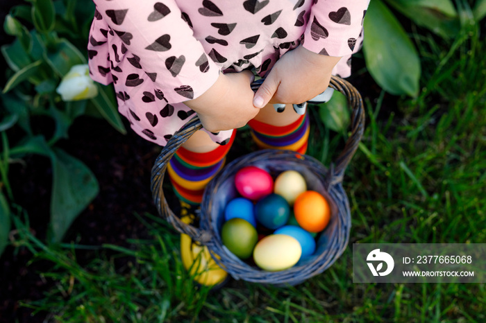 Close-up of legs of toddler girl with colorful stockings and shoes and basket with colored eggs. Child having fun with traditional Easter eggs hunt, outdoors. Celebration of christian holiday.