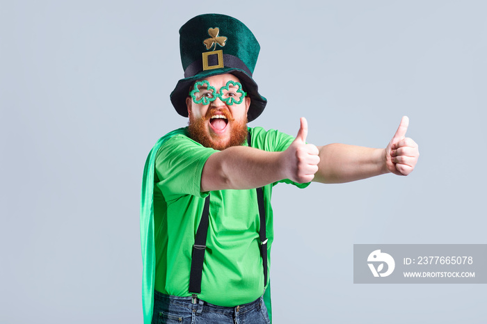 A fat man with a beard in St. Patrick’s suit is smiling with a mug of beer on a gray background.