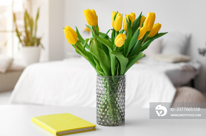 Vase with tulips and book on table in light room