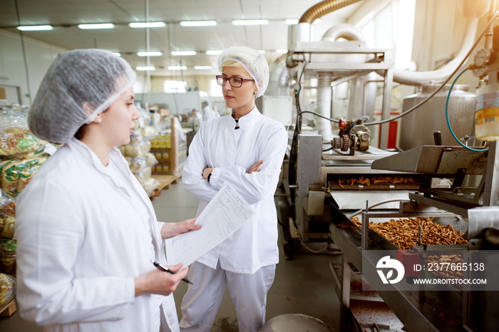 Focused dedicated tired female employee giving rapport in food production factory to her worried and annoyed superior while holding statistic notes and wearing sterile cloths.