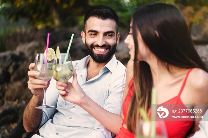 Couple at terrace garden party toasting clinking cocktail glasses and looking in each other eyes - sweethearts sitting outdoors on a couch having fun in a warm summer night