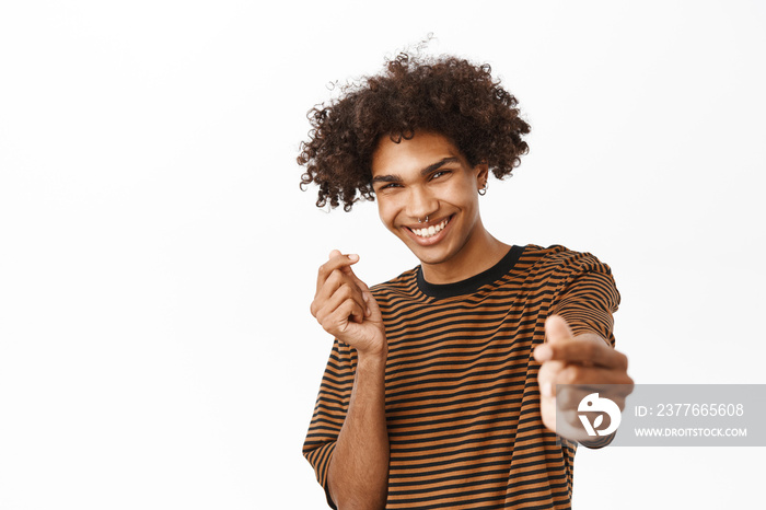 Close up of handsome curly guy shows finger hearts and smiling cute at camera, standing over white background