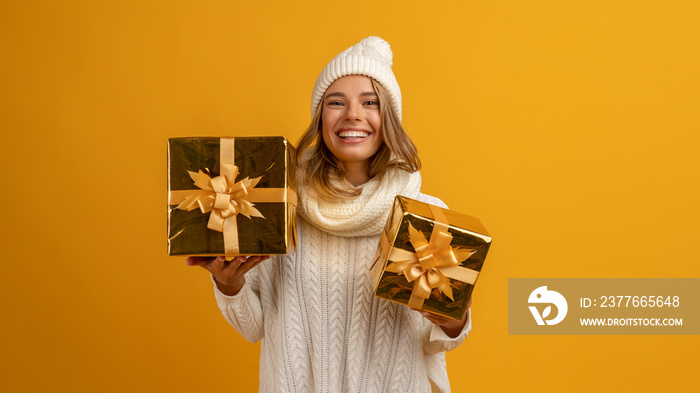 young smiling happy pretty woman holding golden present boxes celebrating new year, christmas gifts, wearing white knitted sweater, scarf and hat, winter fashion trend, posing on yellow background