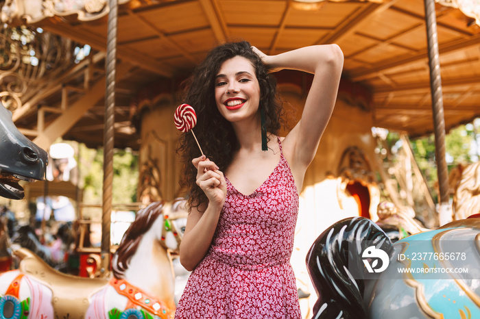 Beautiful lady with dark curly hair in dress standing with lolly pop candy in hand while happily looking in camera with carousel on background